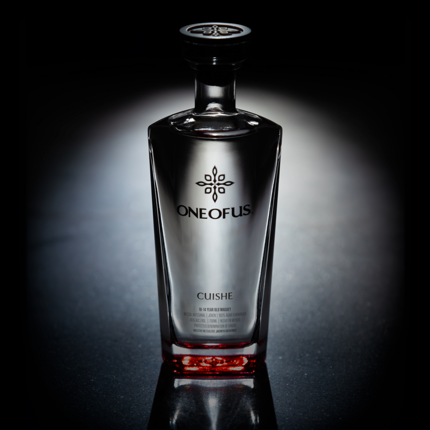 A bottle of ONE OF US mezcal with a clear glass and red label, set against a dark background.