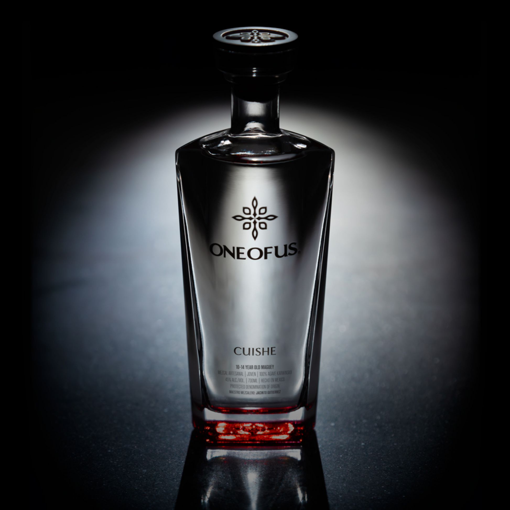 A bottle of ONE OF US mezcal with a clear glass and red label, set against a dark background.